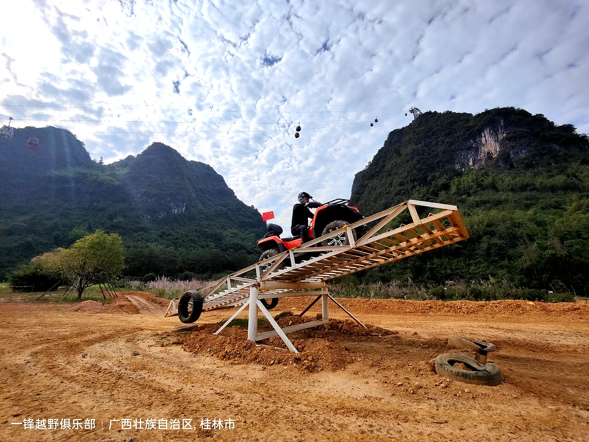 ATV rider navigating a wooden ramp obstacle with karst mountains and cable car in the background, Guangxi China