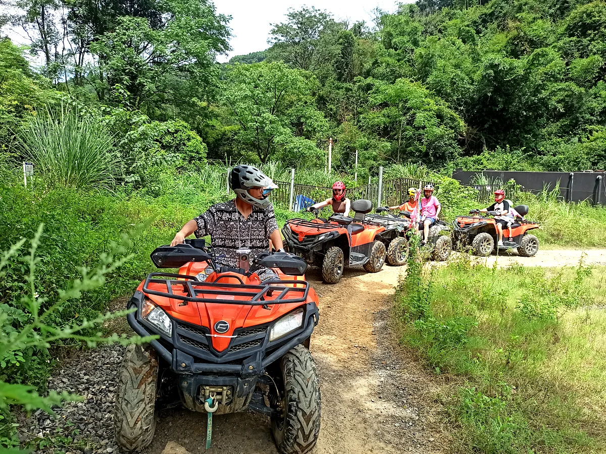 Group of five helmeted riders on orange ATVs driving along an outdoor dirt trail surrounded by green vegetation, China