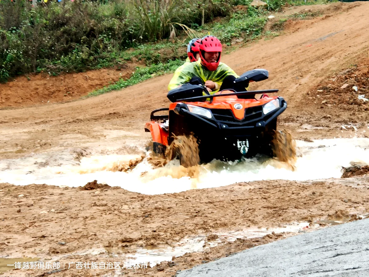 Two riders on an orange ATV splashing through a muddy puddle on a mountain trail, Guangxi China