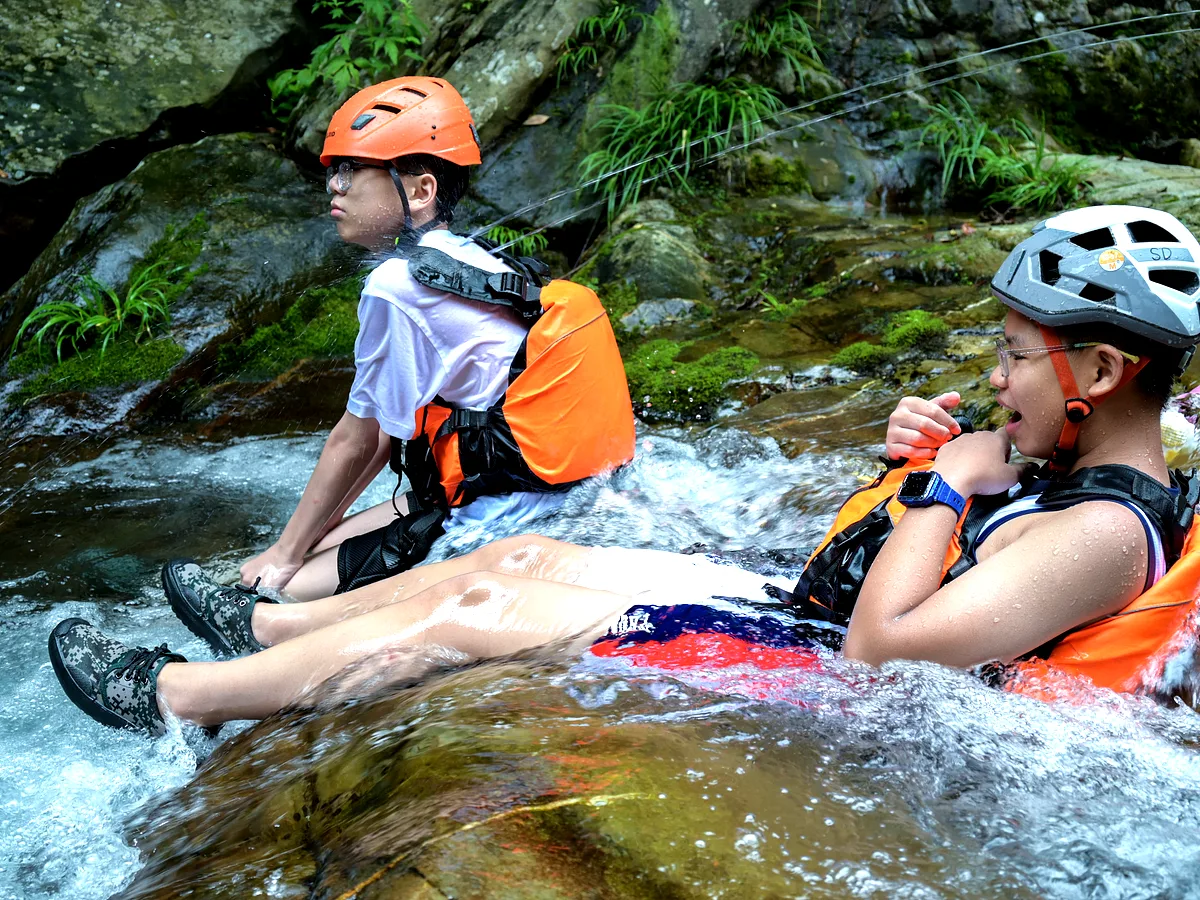 Two children in life jackets and helmets sitting on stream rocks as water flows around them in a canyon, China